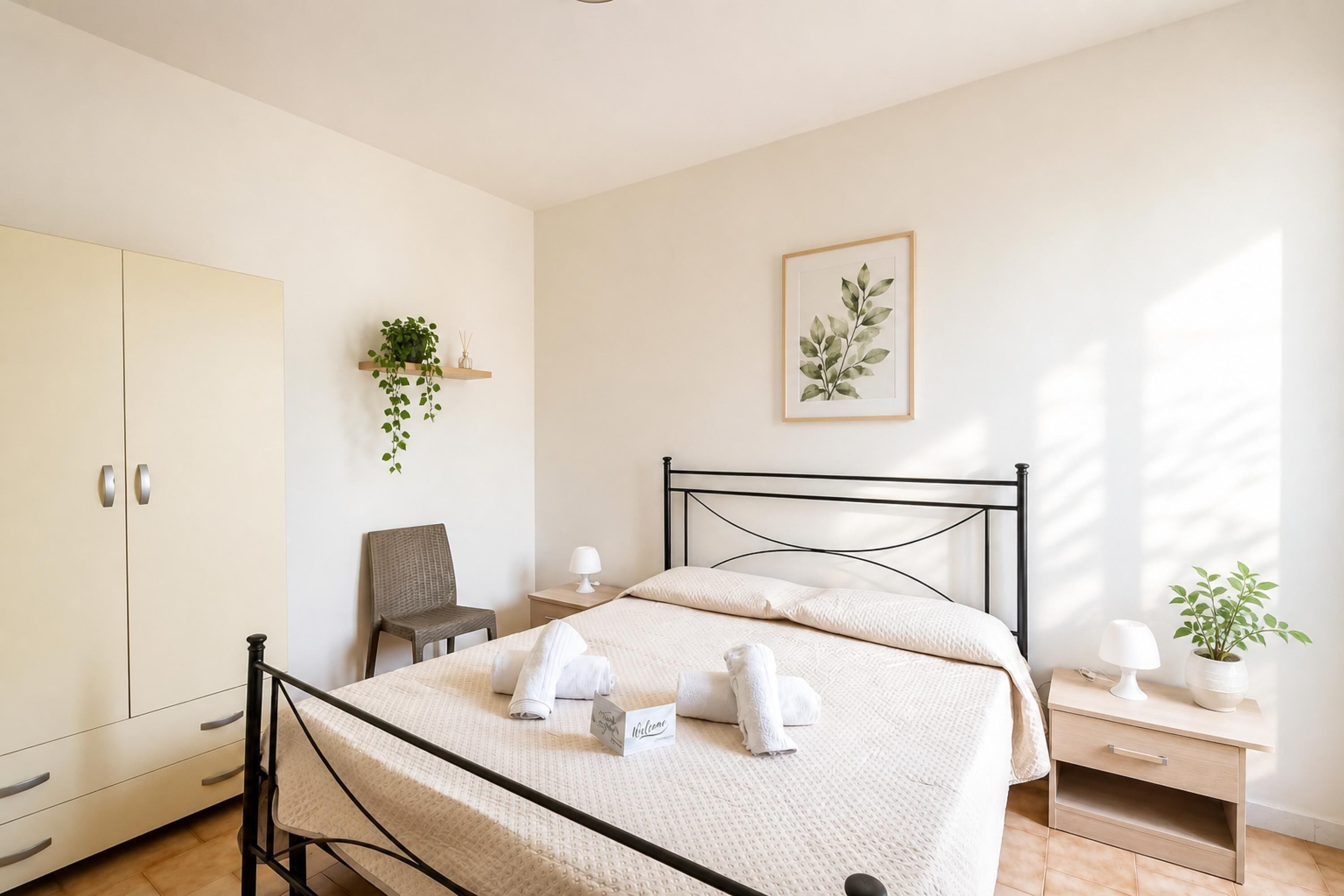 A bedroom with a bed, a wardrobe, a chair, a bedside table, a lamp, and a framed plant on the wall.