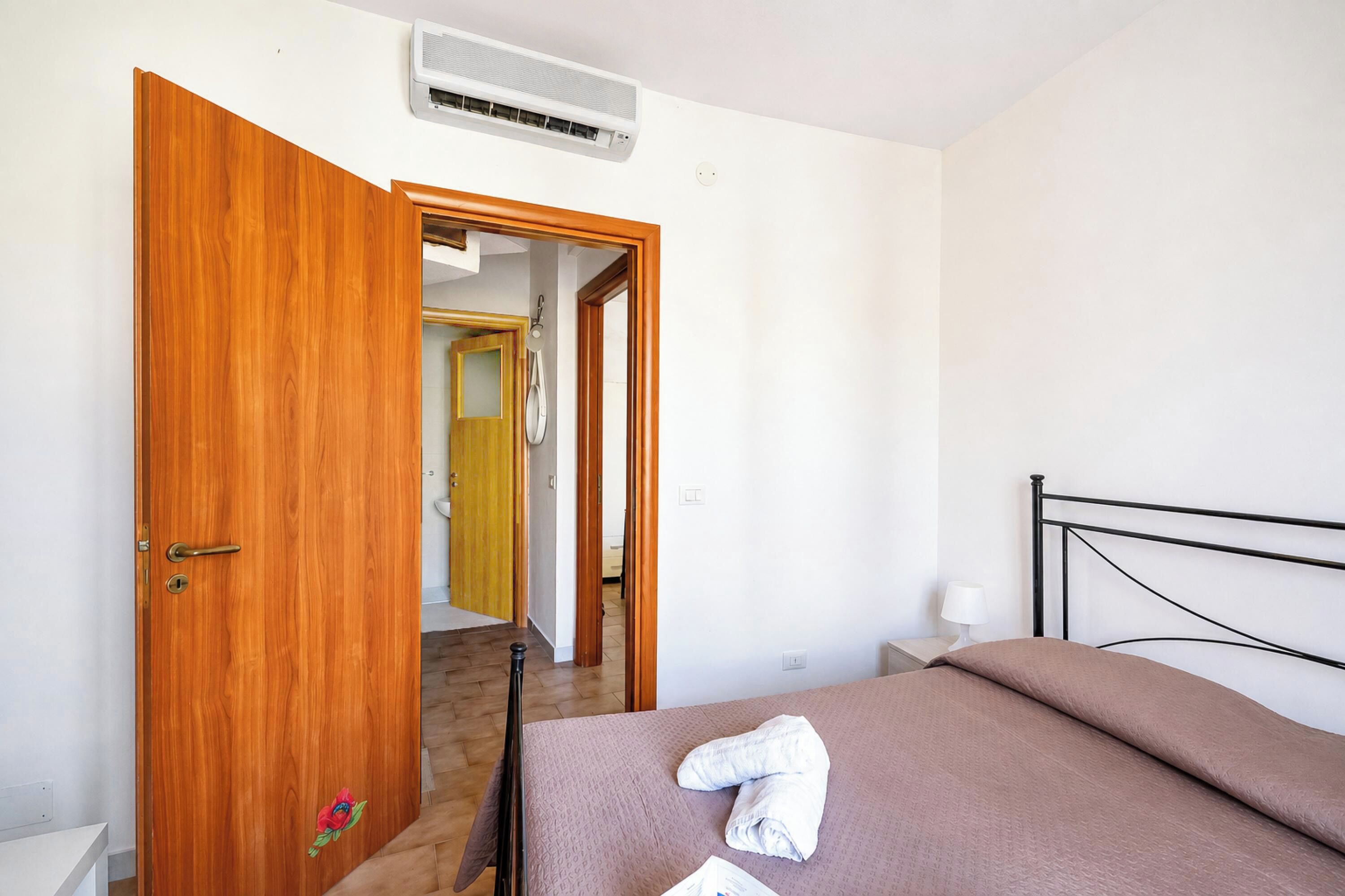 A room with a bed, a wooden door, a bathroom visible through an open door, and a ceiling-mounted air conditioning unit.