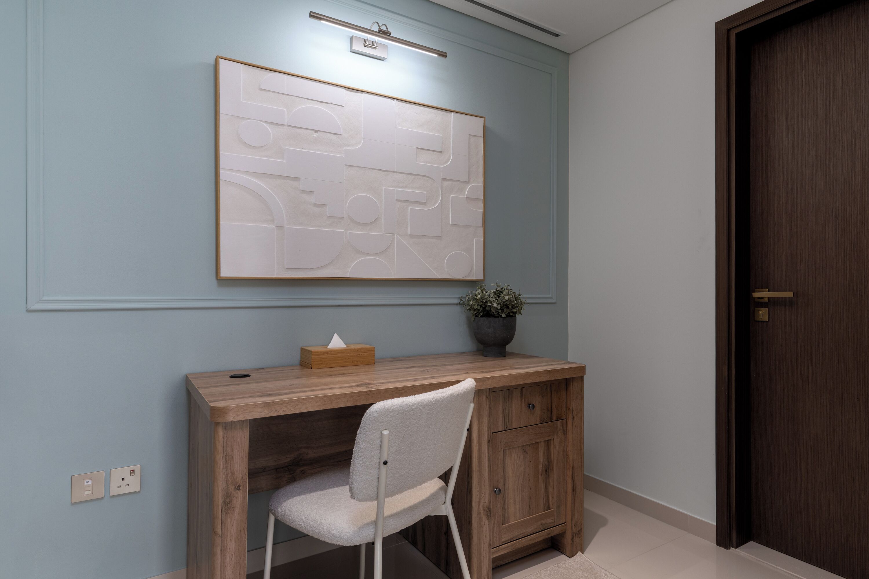 A room with a wooden desk, a chair, a wall-mounted light, a potted plant, and a door.