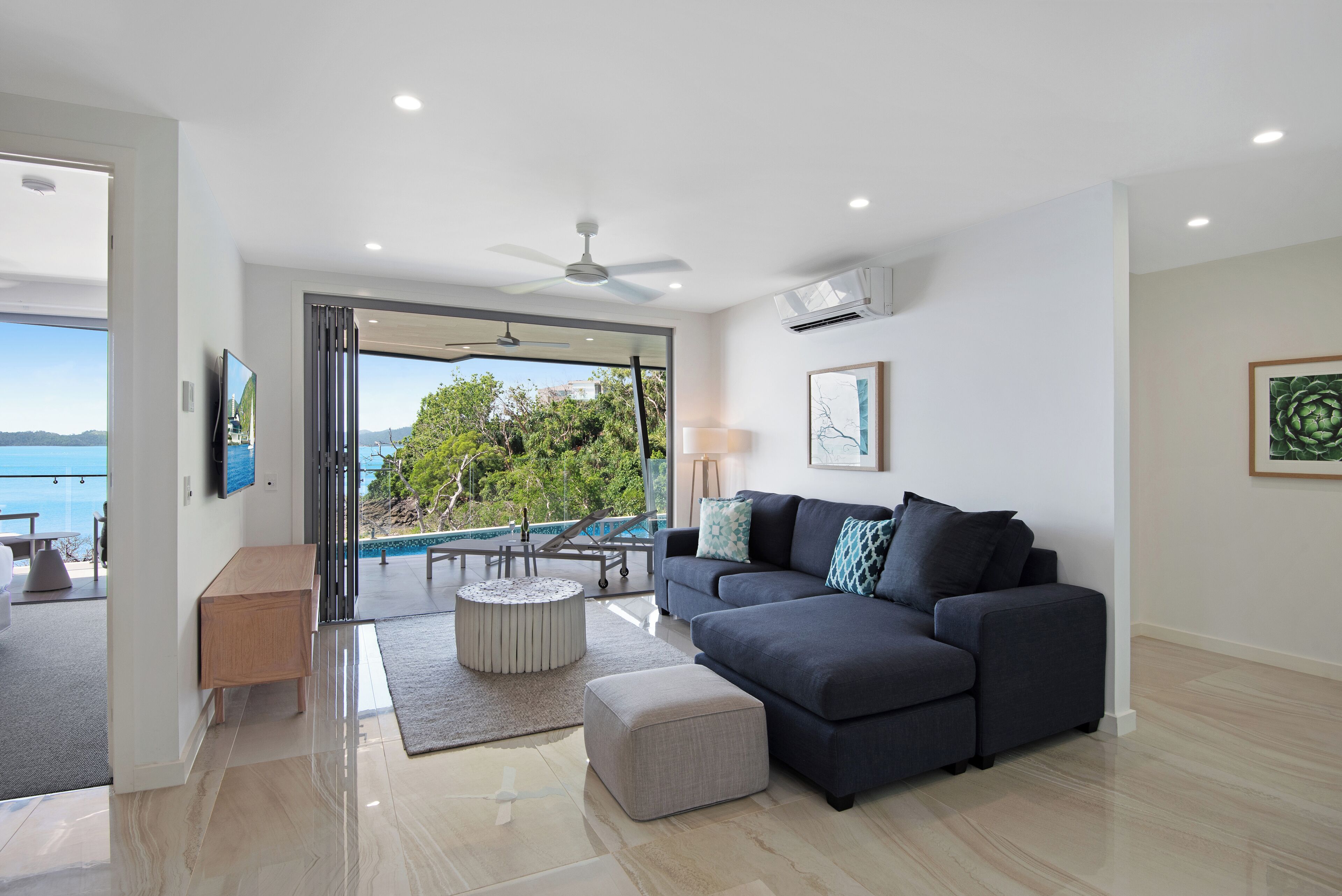 A modern living room with a dark grey sofa, a round ottoman, and a glass door leading to a balcony with a pool view.