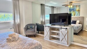 Ein Hotelzimmer mit Bett, Fernseher auf einem Holzständer, Sessel, Schreibtisch, Deckenventilator und einem Gemälde an der Wand.