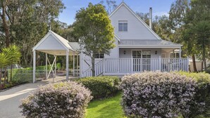 Une maison à deux étages, à l’extérieur blanc, dotée d’un porche et d’un jardin clos, agrémenté d’arbustes en fleurs.