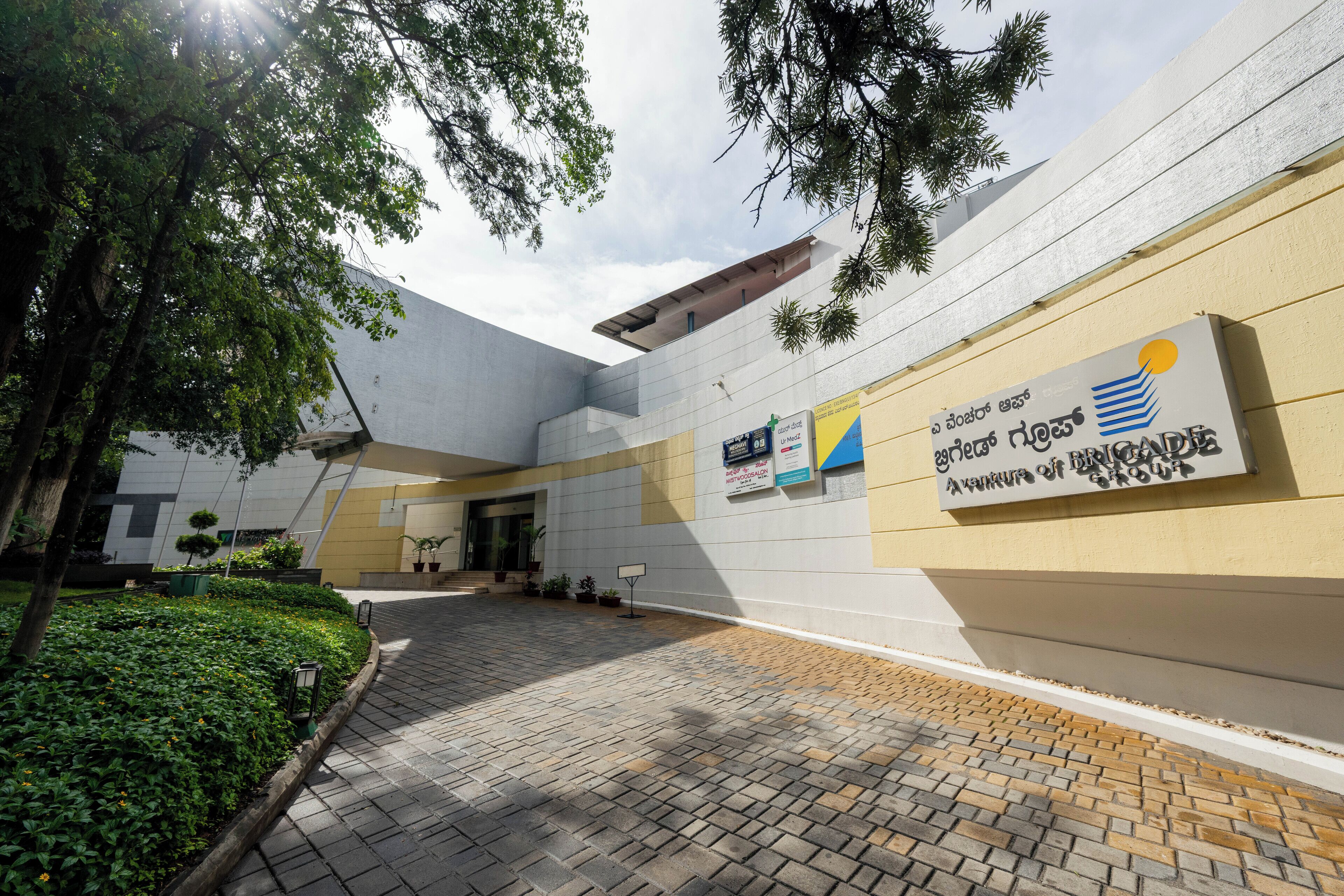A modern building with a paved courtyard, signage in multiple languages, and a mix of greenery and architectural elements.
