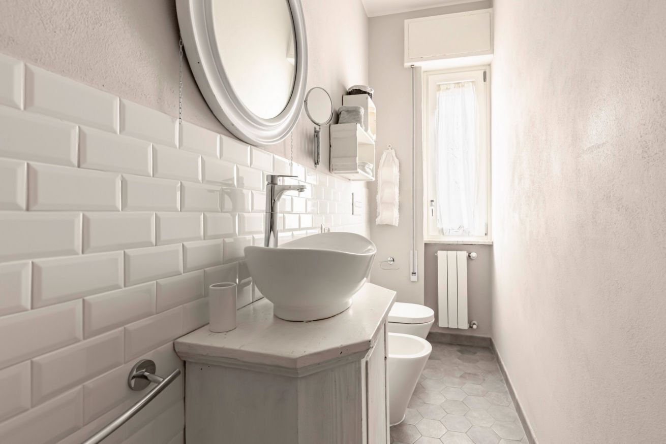 Un bagno con piastrelle bianche tipo metro, un lavandino bianco, uno specchio rotondo e un water.
