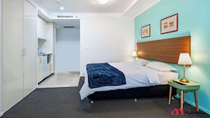 Habitación de hotel con cama, mesita de noche, lámpara y vista al área de la cocina.