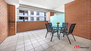 Un balcón con una mesa y sillas de vidrio, un florero y vista a un edificio con múltiples ventanas.