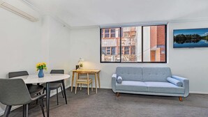 Una habitación con una mesa de comedor, sillas, un sofá gris, una pequeña mesa de madera con una lámpara y una ventana con vista a la ciudad.