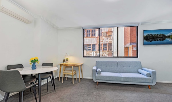 Una habitación con una mesa de comedor, sillas, un sofá gris, una pequeña mesa de madera con una lámpara y una ventana con vista a la ciudad.