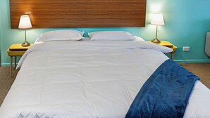 Un lit bien fait, avec une literie blanche et un plaid bleu, flanqué de deux tables de chevet avec des lampes, le tout adossé à un mur de couleur turquoise et agrémenté d’une tête de lit en bois.