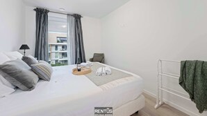 Ein ordentlich bezogenes Bett mit Kissen, eine Nachttischlampe, ein Korb mit einer Flasche und Handtüchern sowie ein Blick auf ein Gebäude durch das Fenster.