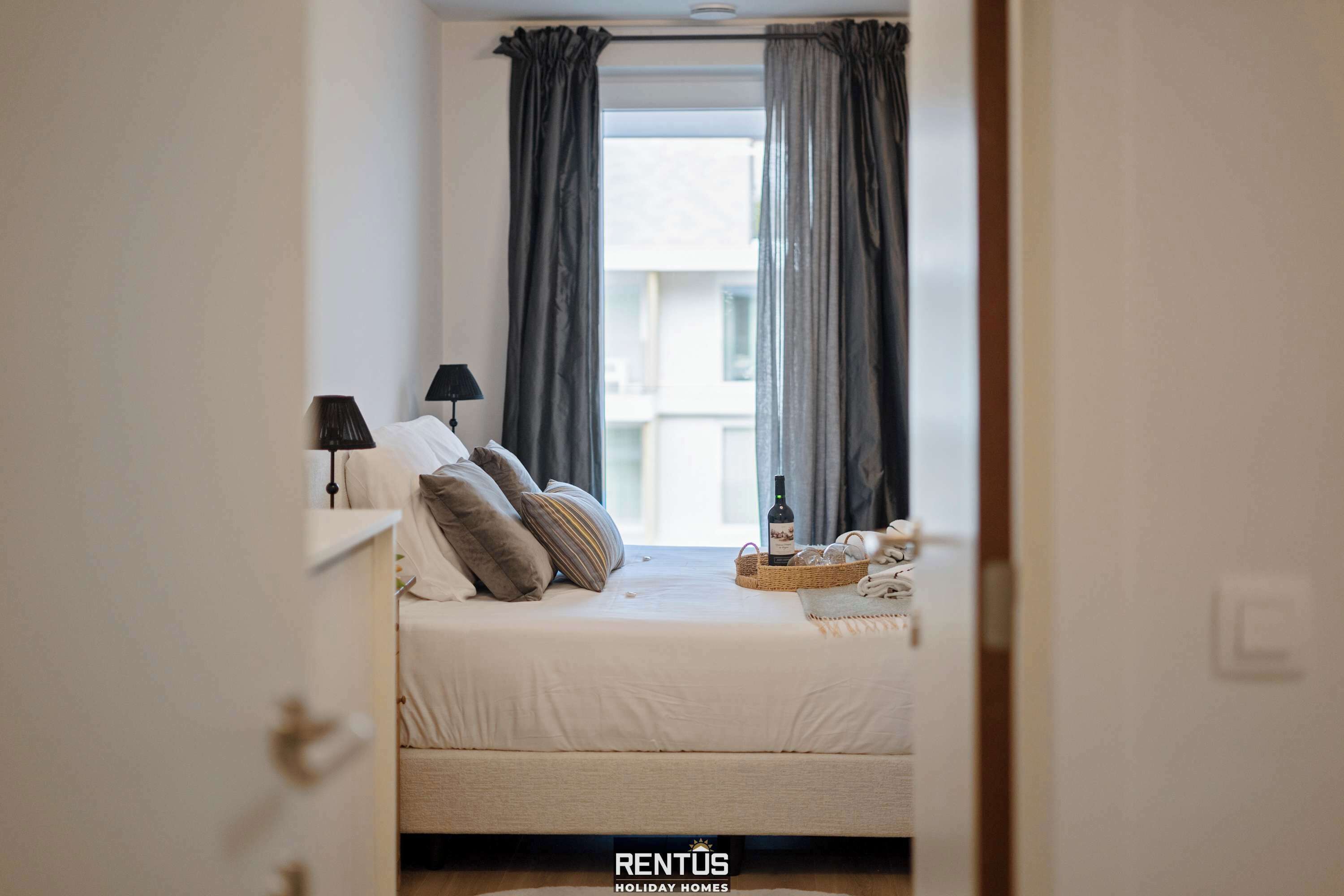A bedroom with a bed, pillows, a lamp, a basket with items, and a window with curtains.