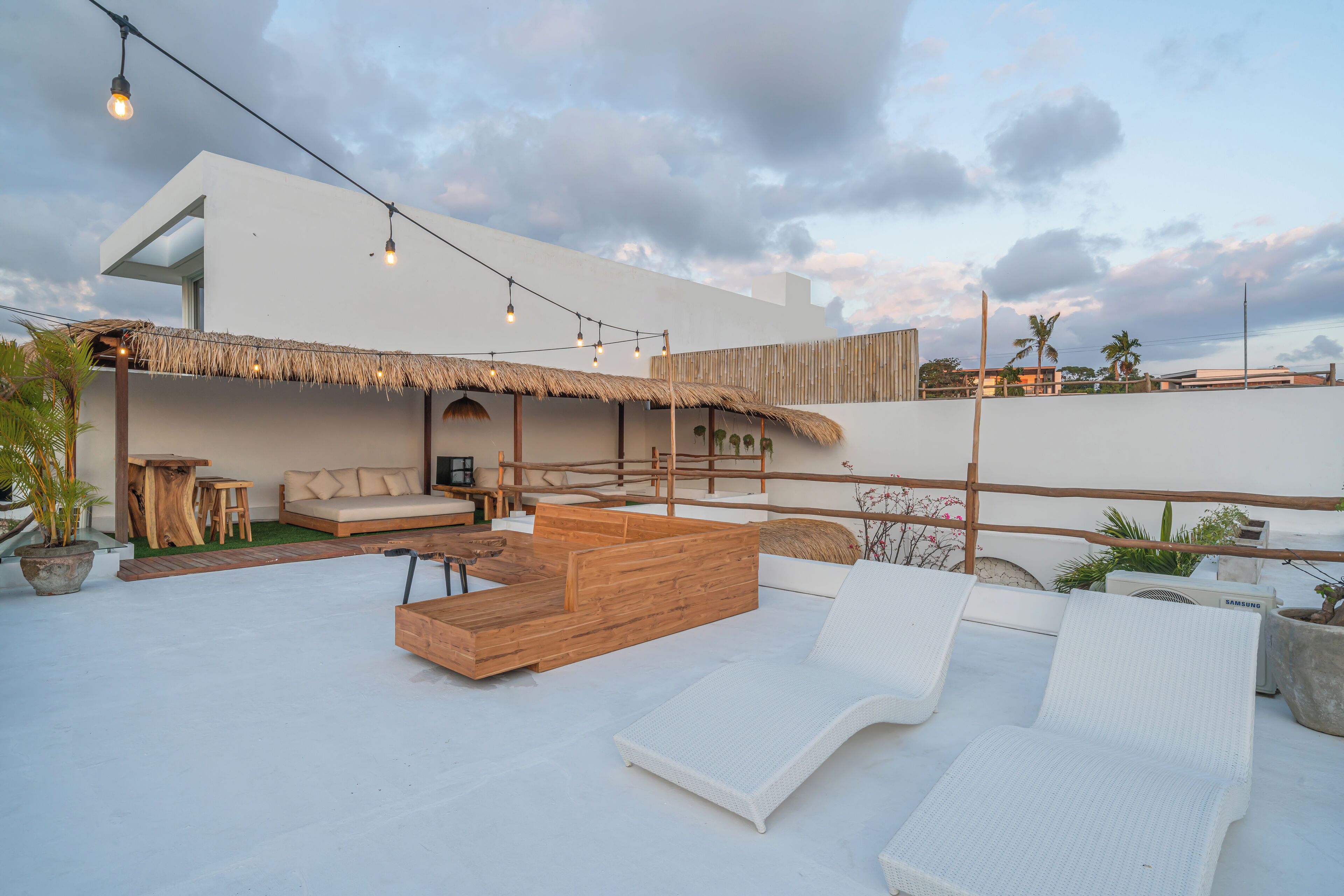 Un espace de détente extérieur avec un banc en bois, des fauteuils blancs et des guirlandes lumineuses.