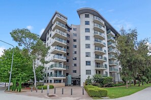 A modern multi-story residential building with balconies, a landscaped entrance, and a clear sky.
