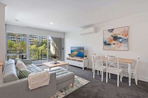 A modern living room with a grey sofa, a dining table with chairs, a flat-screen TV, and a large window with a view of buildings and trees. - Stylish 1-Bed Apartment with Balcony & Pool Access (Turner)