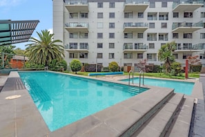 Un moderno edificio de apartamentos con piscina, rodeado de palmeras y jardines bien cuidados.