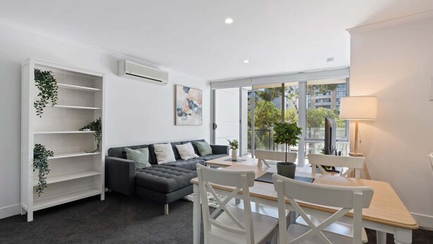 A modern living room with a grey sofa, white chairs, a small table, and a large window offering a view of buildings and trees. - Central Braddon Retreat with Balcony & Pool Access (Turner)