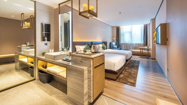 Une chambre d’hôtel moderne avec un grand miroir, une coiffeuse en marbre, un lit avec une tête de lit en bois, une table de chevet et un petit bureau avec une lampe.