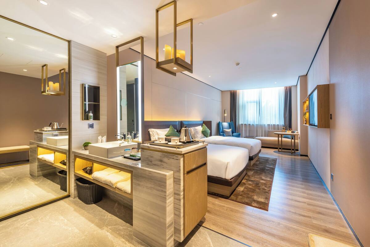 Quarto de hotel moderno com um espelho grande, toucador de mármore, cama com cabeceira de madeira, mesa de cabeceira e pequena escrivaninha com abajur.