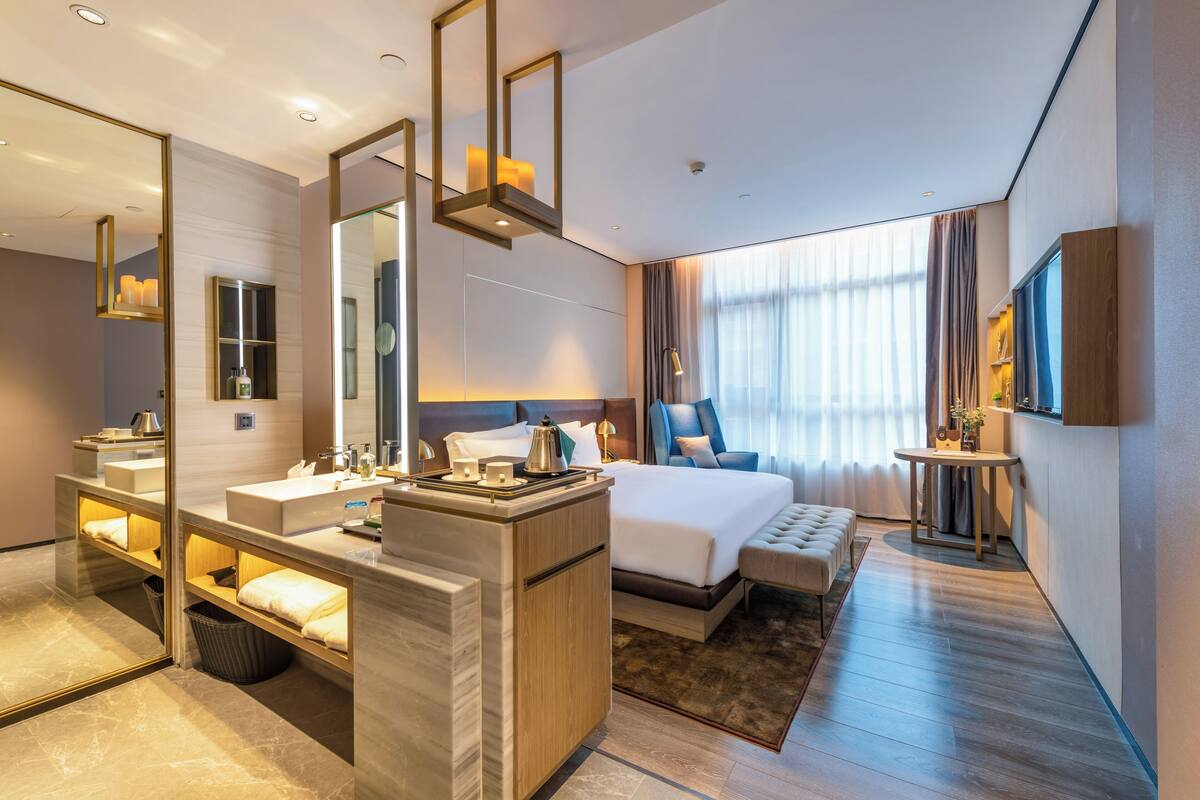 Quarto de hotel moderno com uma cama grande, banheiro separado com toucador, área de estar e televisão fixada na parede.
