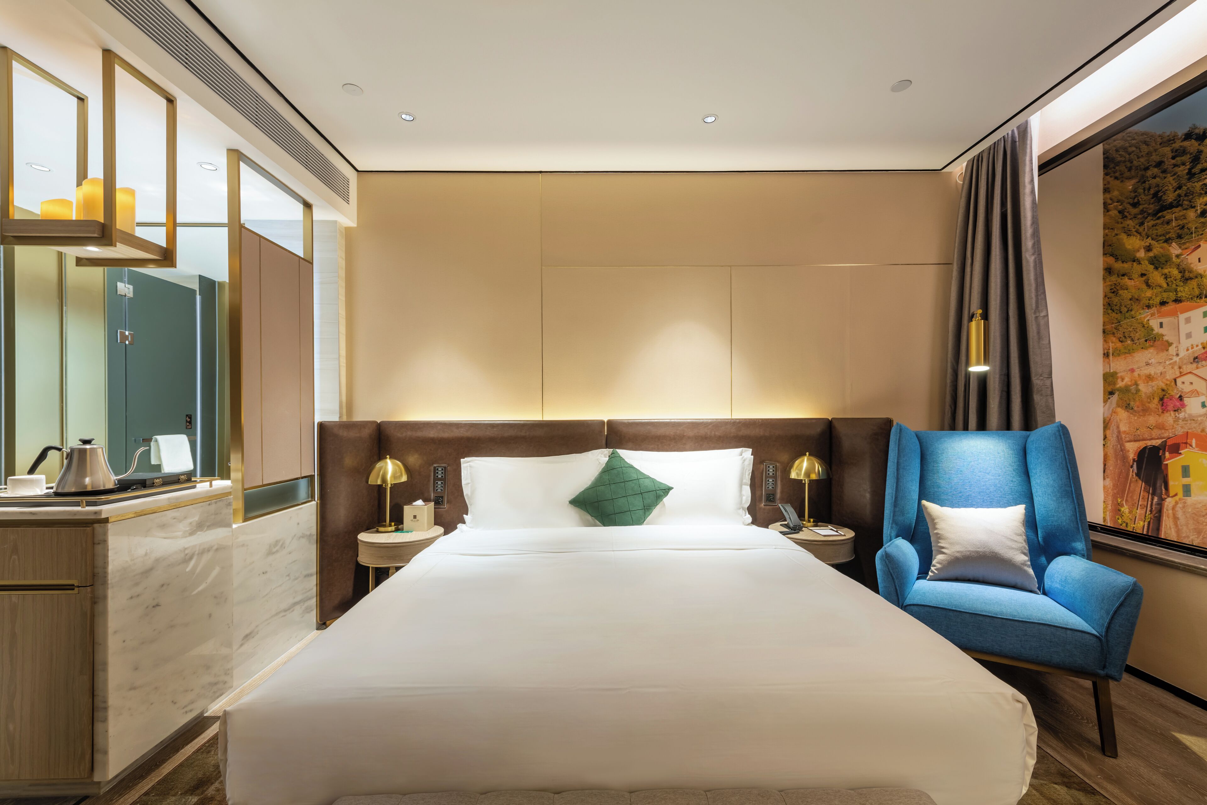 Ein Hotelzimmer mit einem großen Bett, einem blauen Sessel, einem Schreibtisch mit Lampe und einem großen Fenster mit Vorhängen.