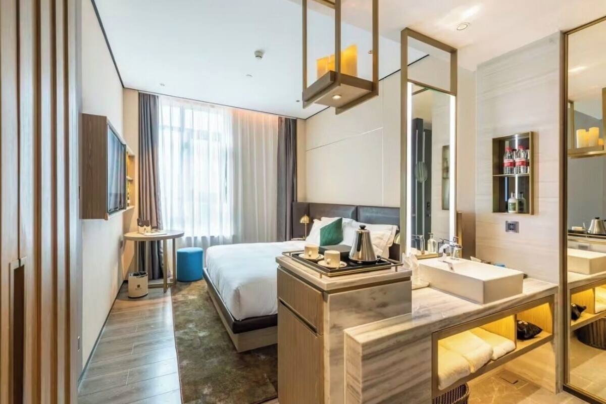 Quarto de hotel moderno com uma cama grande, uma mesinha de cabeceira com luminária, televisão integrada, um espelho grande e banheiro com pia e espelho.