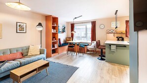 Ein modernes Wohnzimmer mit einer Couch, einem Couchtisch und einem Essbereich mit Stühlen und einem Tisch. Es gibt ein Bücherregal, ein Fenster mit Vorhängen sowie eine Küchenzeile mit Arbeitsfläche und Spüle.
