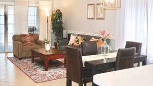 A living room with a sofa, coffee table, dining table, chairs, a fireplace, and a large window with curtains. - Coralbell Villa - Near UNCC & PNC Pavilion, Pools, Grill, Spacious, 15 Mins to Uptown, King Suite! (Charlotte)