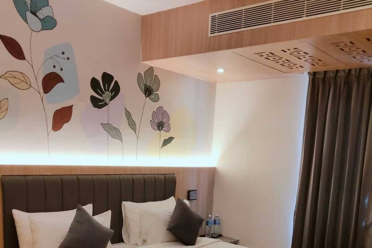 A hotel room with a large bed, a headboard, a wall with floral artwork, a ceiling fan, and a window with curtains.