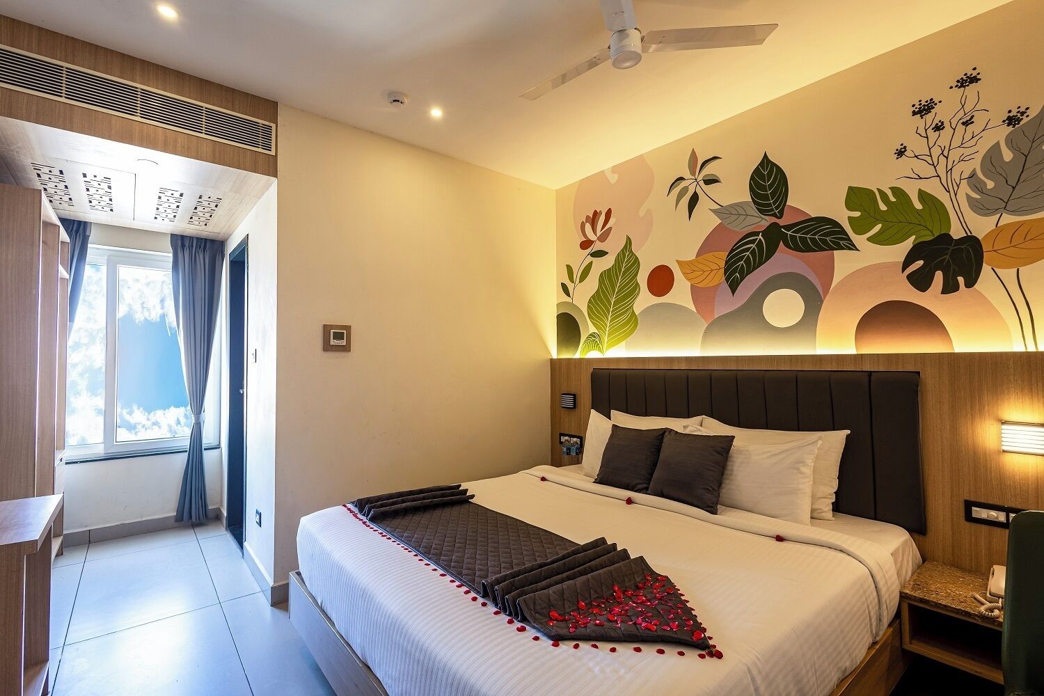 A hotel room with a large bed, a ceiling fan, a wall with a leaf pattern, and a window with curtains.