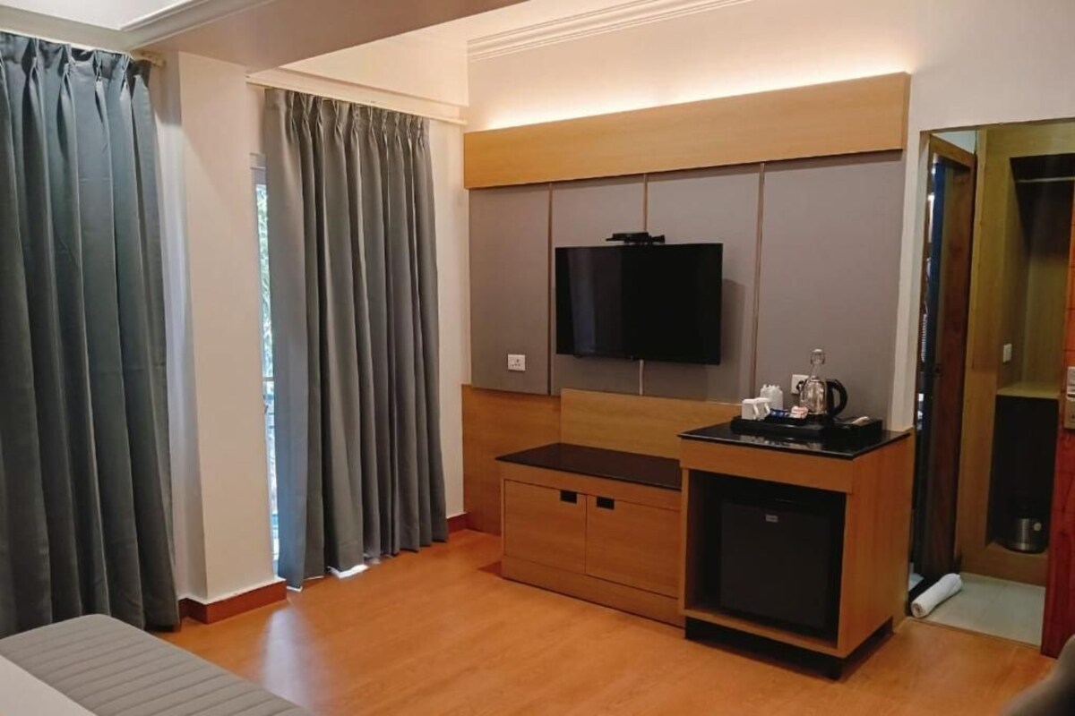 A hotel room with a bed, a TV mounted on the wall, a wooden cabinet, and a small kitchenette area.