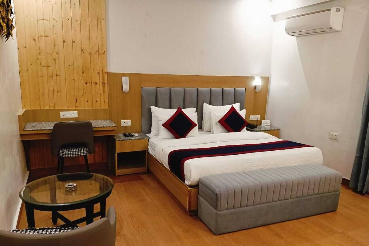 A hotel room with a bed, a small desk, a chair, a glass table, and an air conditioning unit.
