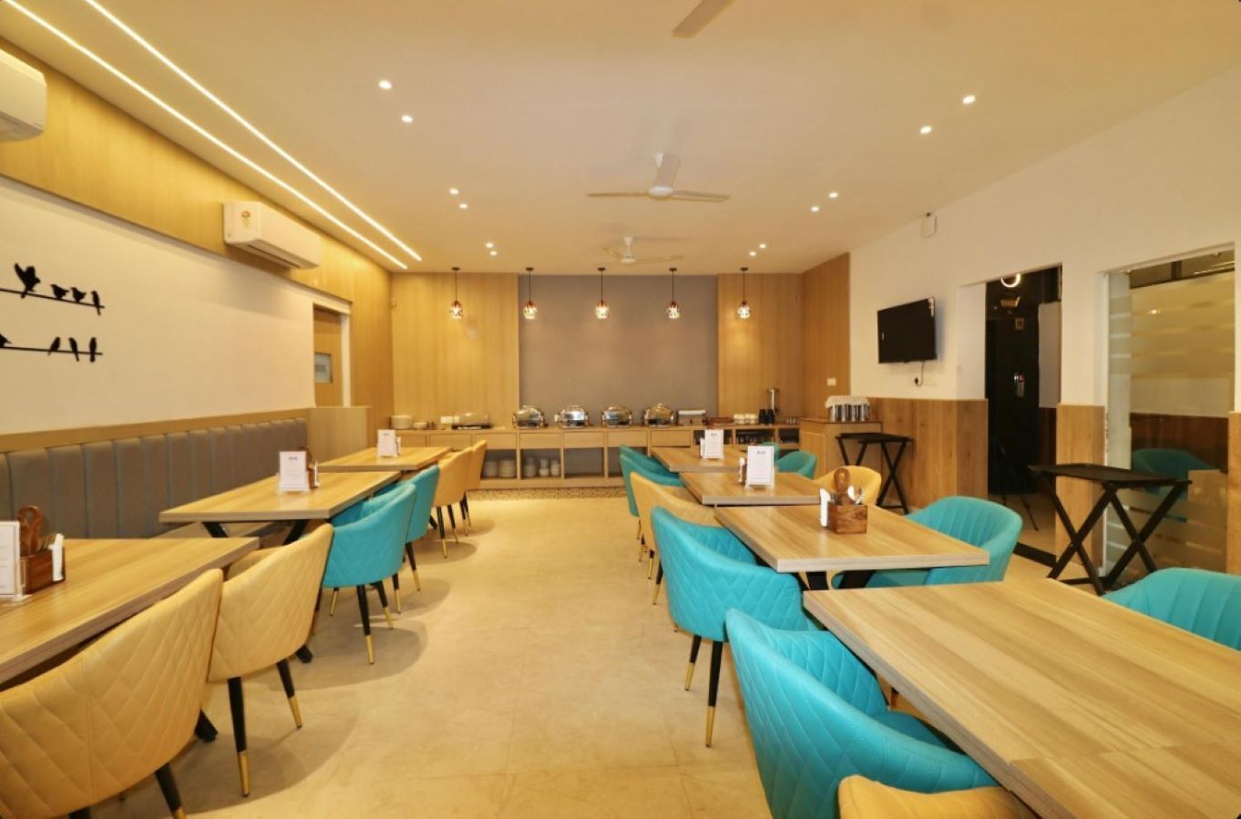 Un'area pranzo moderna con tavoli di legno e sedie color petrolio, una cucina sullo sfondo e un televisore a schermo piatto montato a parete.
