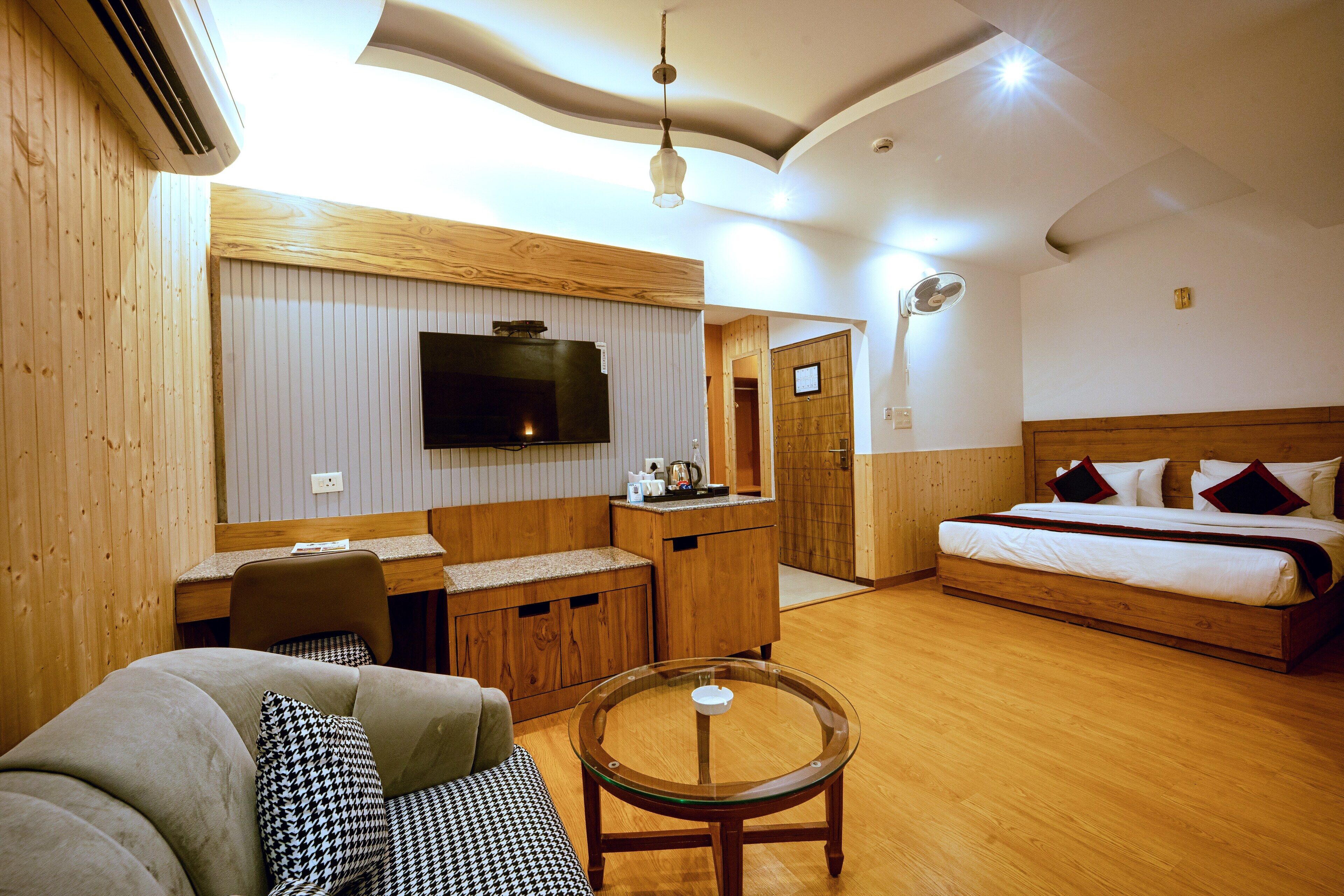 Une chambre d’hôtel avec un plancher en bois, un lit doté d’une tête de lit en bois, un canapé avec des coussins à motifs, une table en verre, un téléviseur fixé au mur et un petit coin cuisine.
