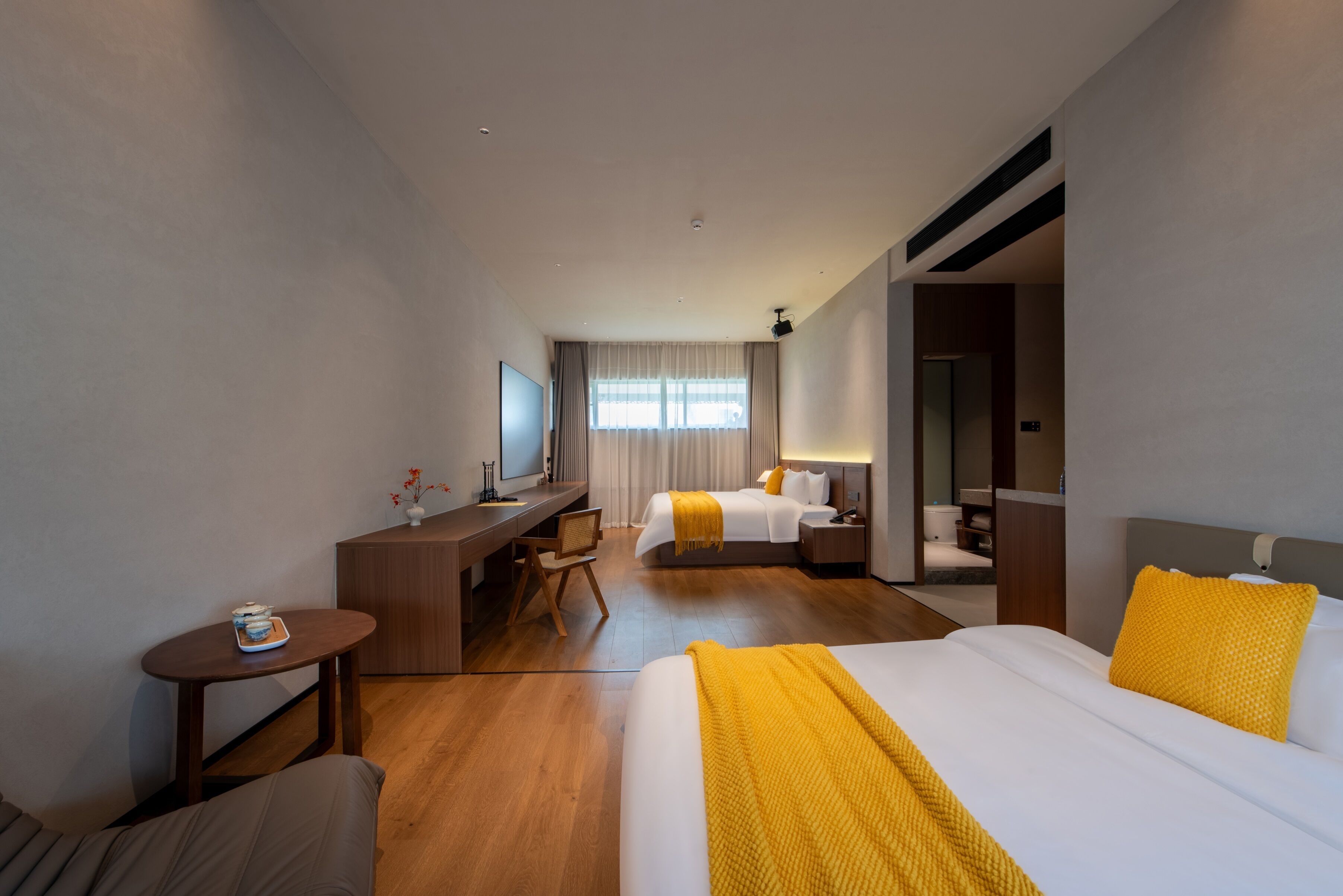Quarto de hotel com uma cama grande, uma cama menor, uma escrivaninha com cadeira, televisão e um banheiro visível através de uma porta aberta.