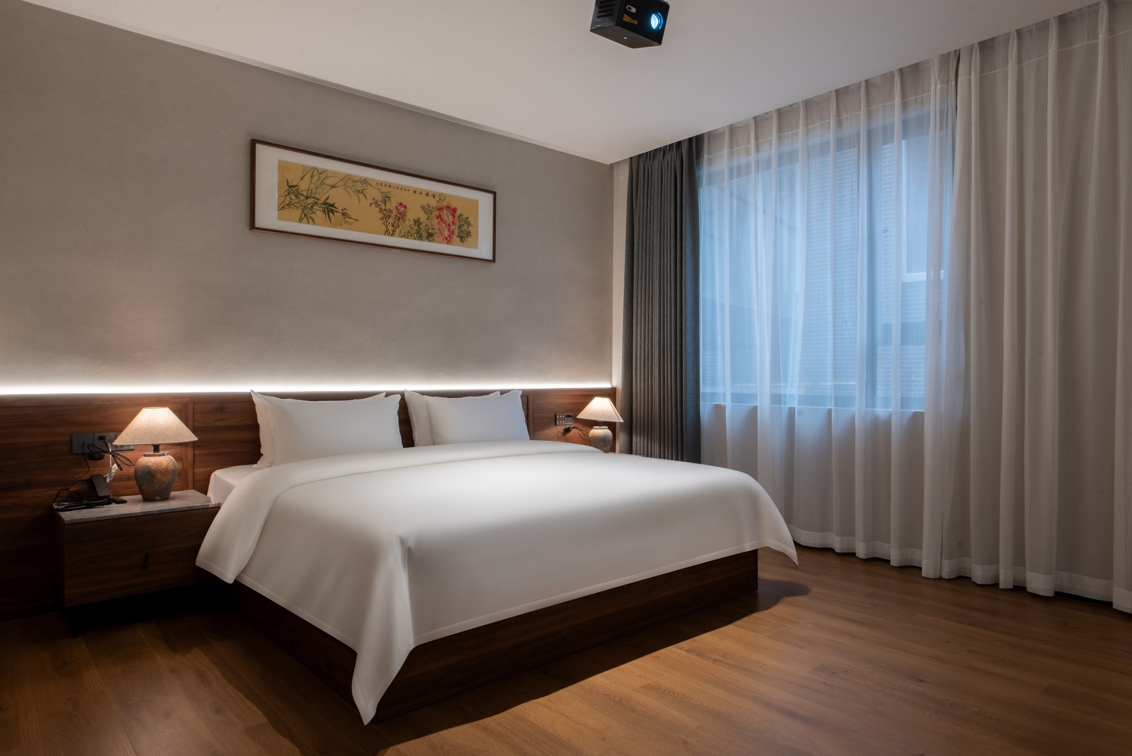 A modern hotel room with a large bed, wooden headboard, two bedside lamps, a nightstand, a framed artwork on the wall, and a large window with sheer curtains.