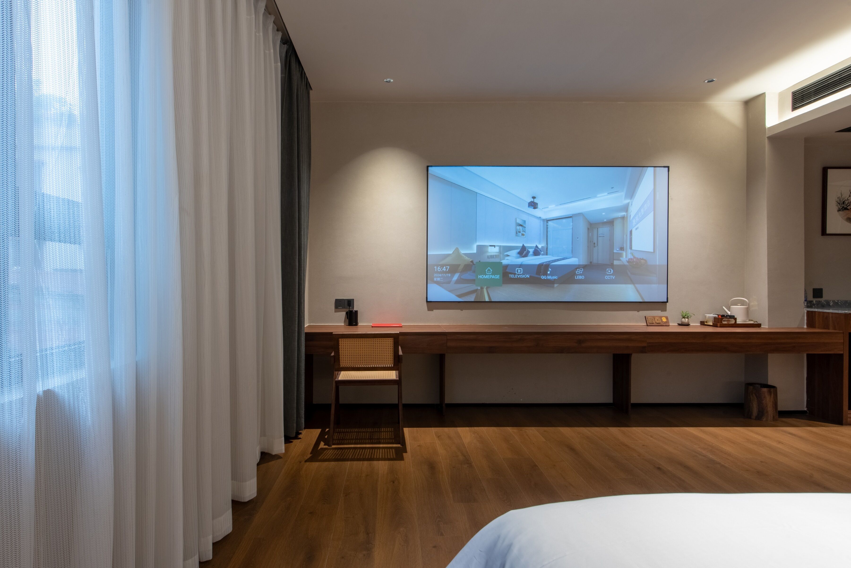 A hotel room with a large TV screen displaying a room layout, a wooden desk, a chair, and a bed.