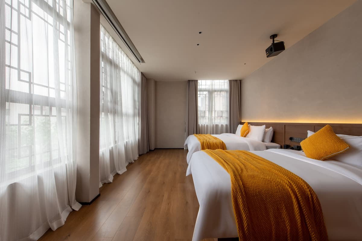 Ein Hotelzimmer mit zwei Betten, einem großen Fenster mit durchsichtigen Vorhängen, Holzboden und einem Deckenprojektor.
