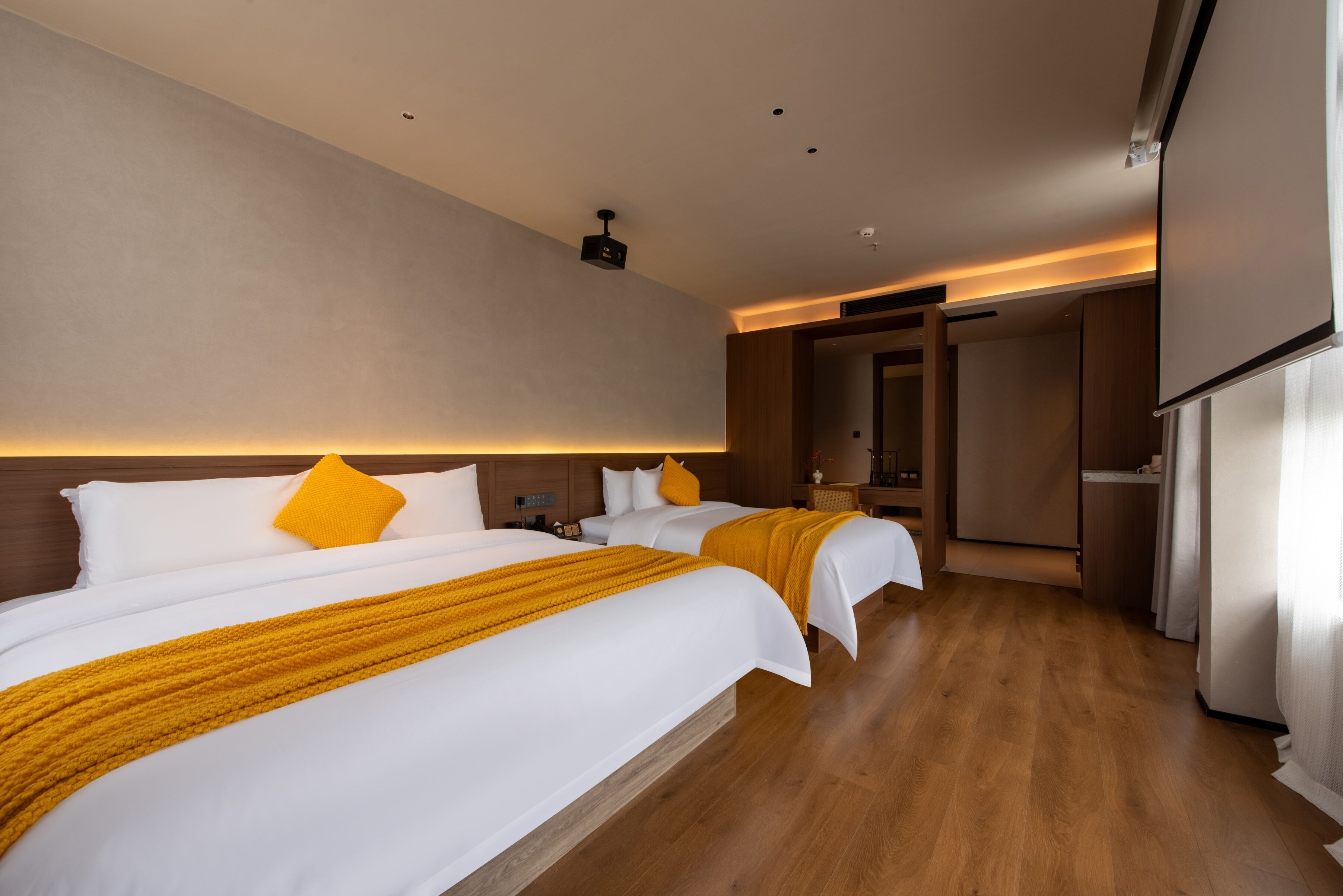 Habitación de hotel con dos camas, piso de madera, una lámpara de techo moderna y una puerta que da a otra habitación.