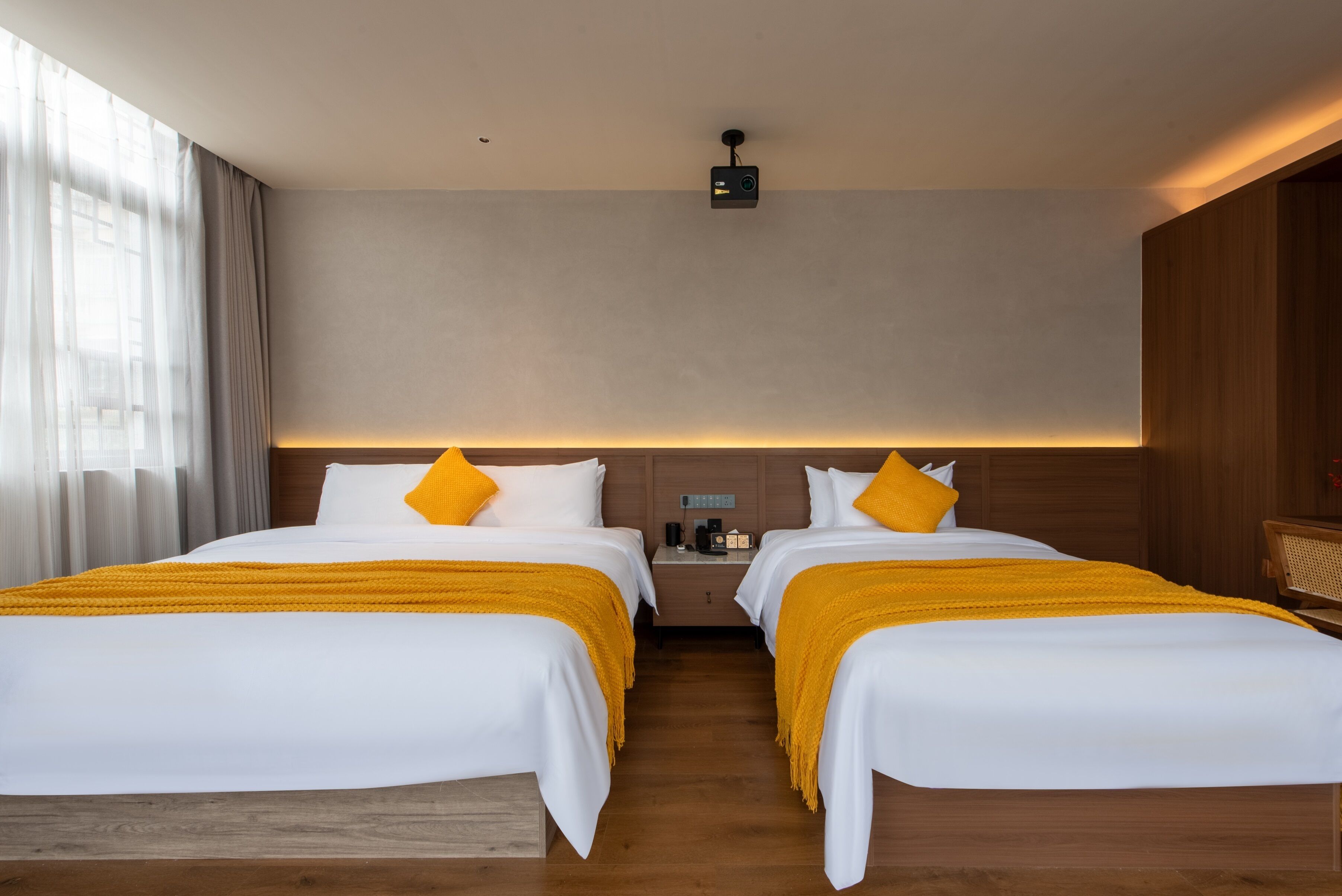 A hotel room with two beds, each with white bedding and yellow throw pillows. There is a nightstand with a lamp and a small table between the beds. The room has a wooden floor and a large window with sheer curtains.