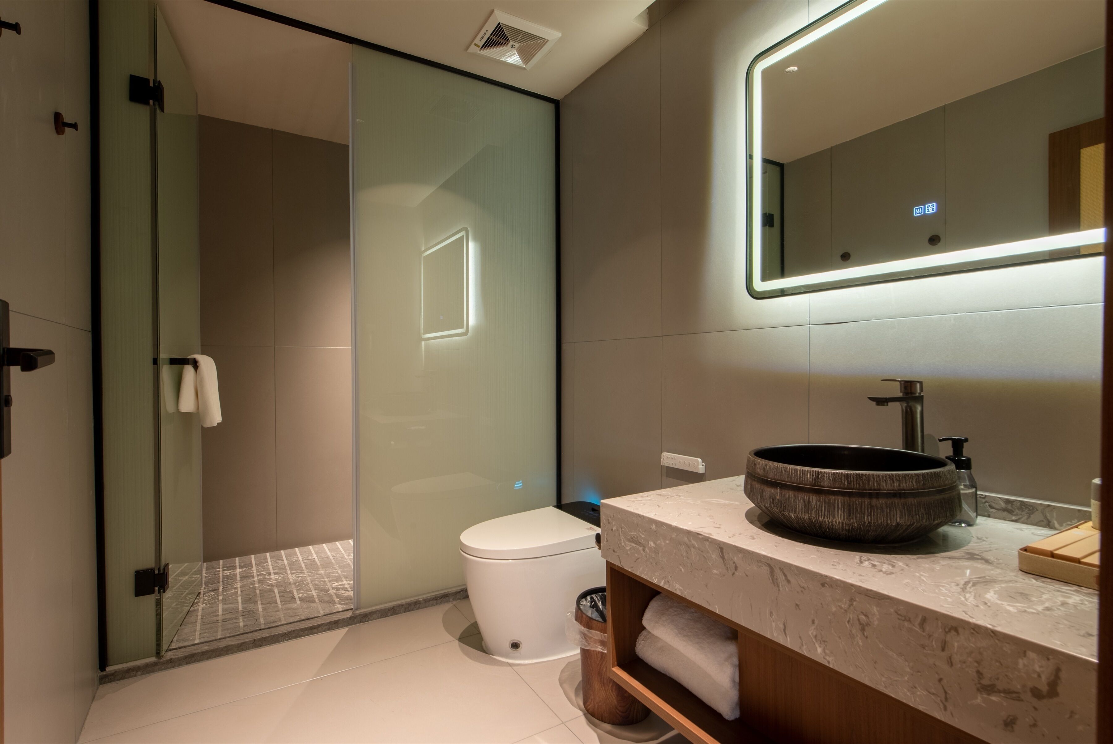 A modern bathroom with a glass shower enclosure, a white toilet, a marble sink, and a mirror with integrated lighting.