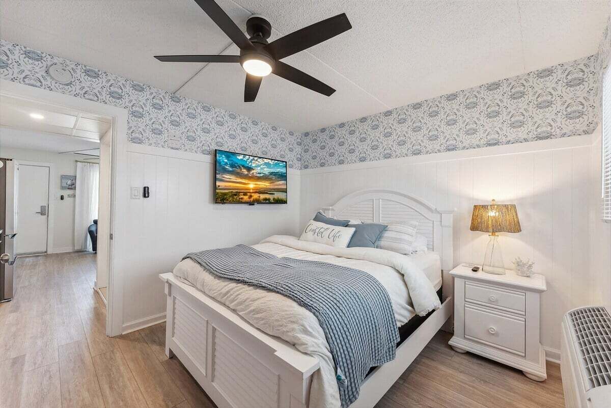 Ein Schlafzimmer mit Bett, Nachttisch, Lampe, Deckenventilator und einem Wandbild.