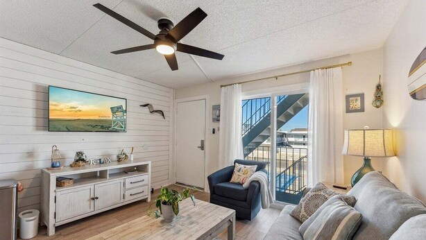 Ein Wohnzimmer mit Deckenventilator, einem Fernseher, der eine Strandansicht zeigt, einem Sofa, einem Couchtisch und einer Treppe, die zu einer oberen Ebene führt.