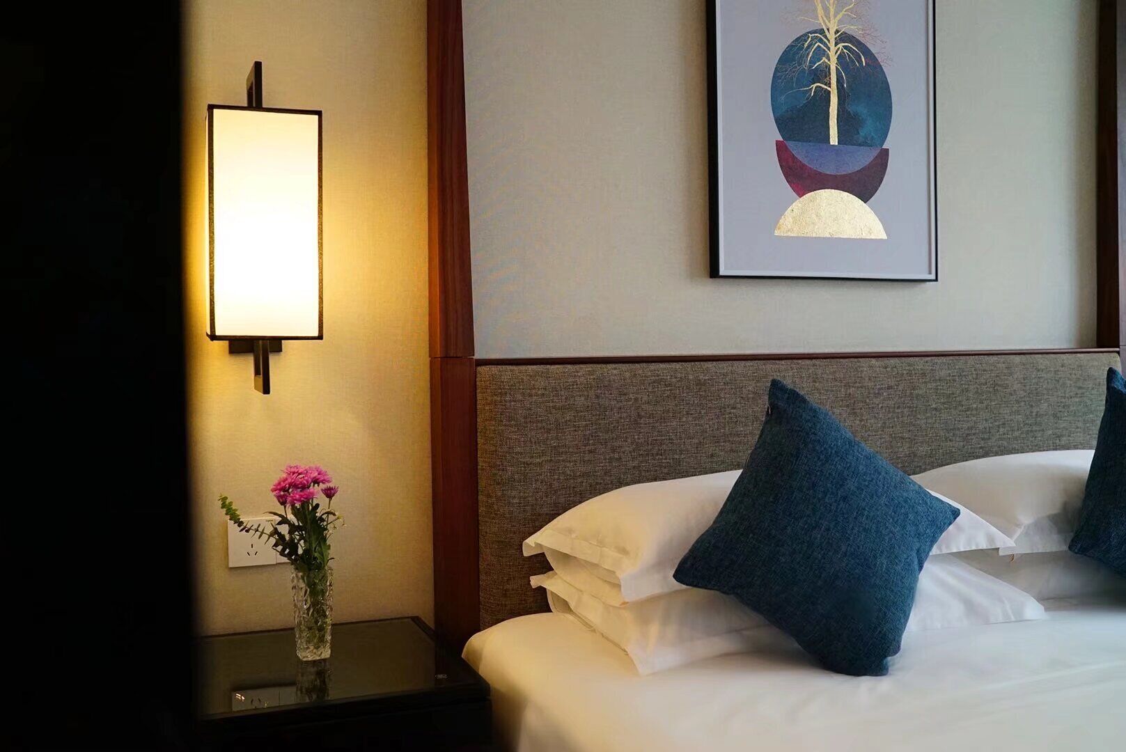 Habitación de hotel con cama, almohadas, mesita de noche con un florero y una lámpara de pared, además de una obra de arte enmarcada.