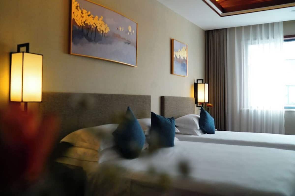 Ein Hotelzimmer mit zwei Betten, jeweils mit Kopfstütze und Kissen. An beiden Seiten der Betten befinden sich Wandleuchten.