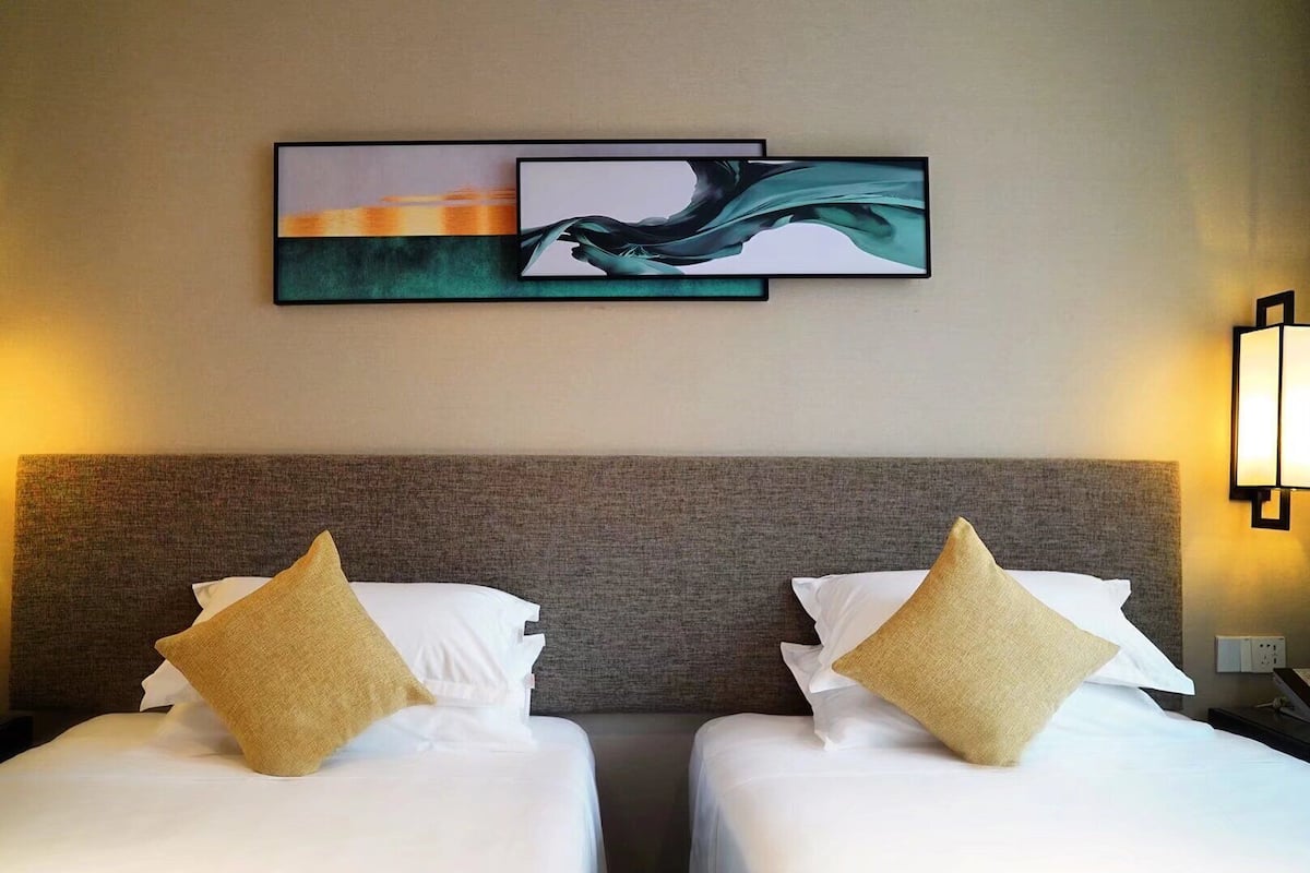 Ein Hotelzimmer mit zwei Betten, jedes mit einer weißen Matratze und einem goldenfarbenen Kissen. Über den Betten hängt ein gerahmtes abstraktes Gemälde.