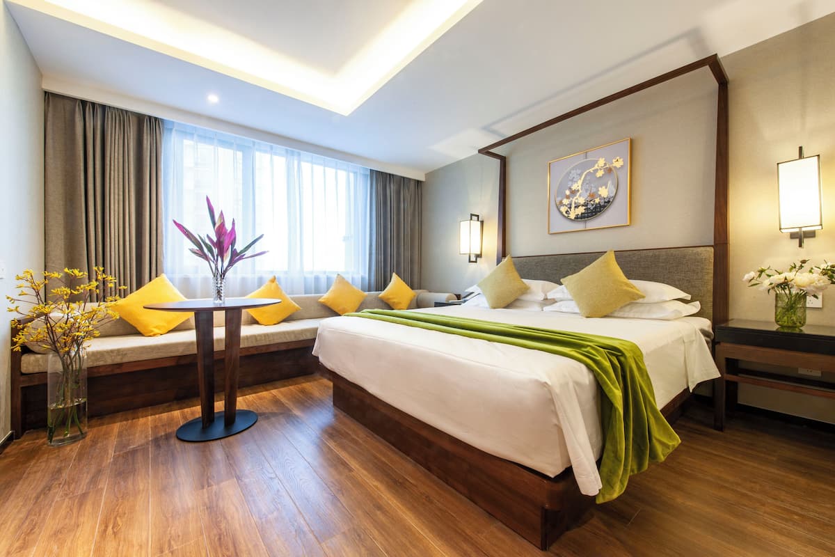 Ein Hotelzimmer mit einem großen Bett, einem runden Tisch, einer Couch mit gelben Kissen, einer Vase mit Blumen und einem Gemälde an der Wand.
