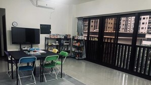 Une pièce dotée d’une grande fenêtre, d’un téléviseur à écran plat, d’une table avec des chaises et d’étagères remplies de livres et de fournitures.