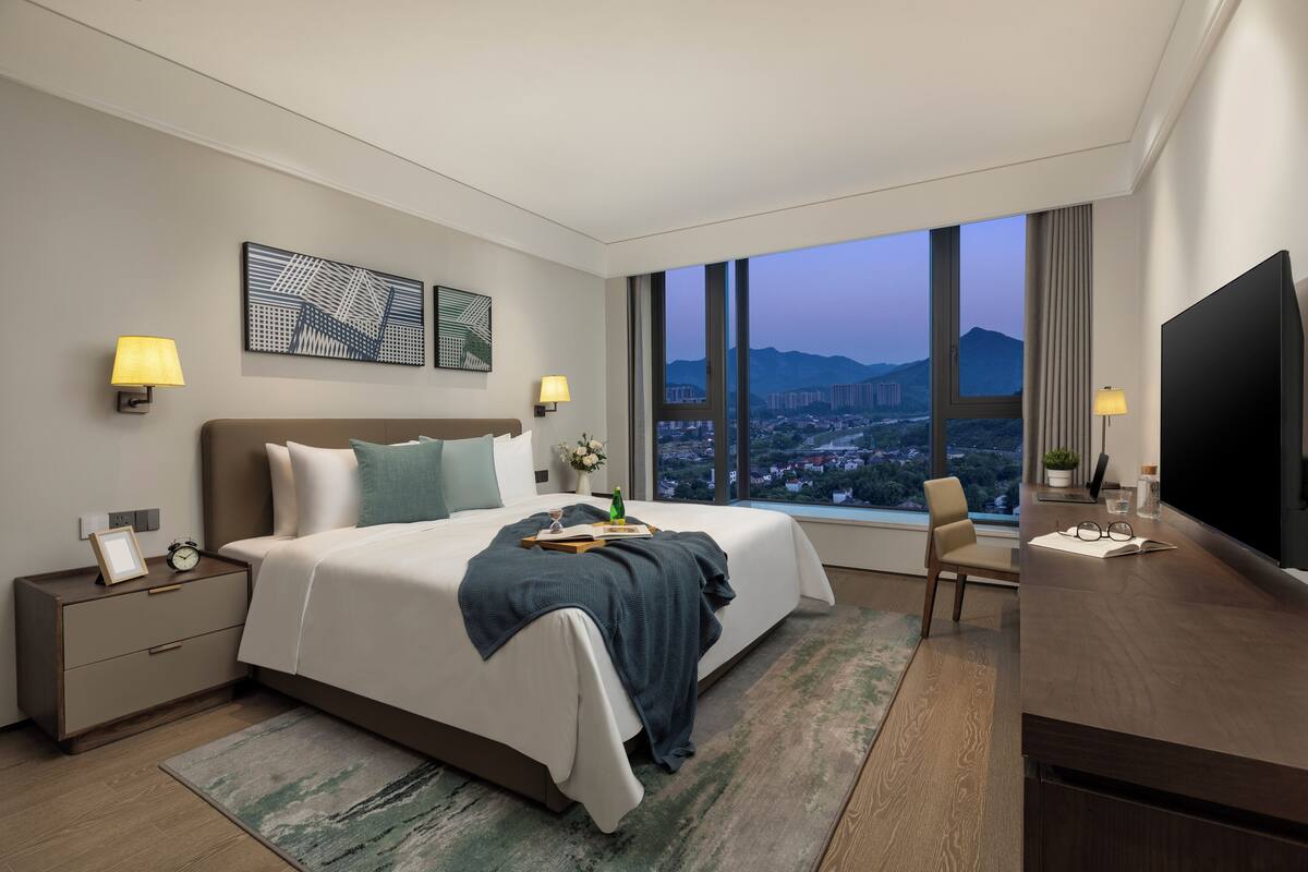 Ein modernes Hotelzimmer mit einem großen Bett, Nachttischlampen, einem Flachbildfernseher und Blick auf eine Stadtlandschaft durch große Fenster.