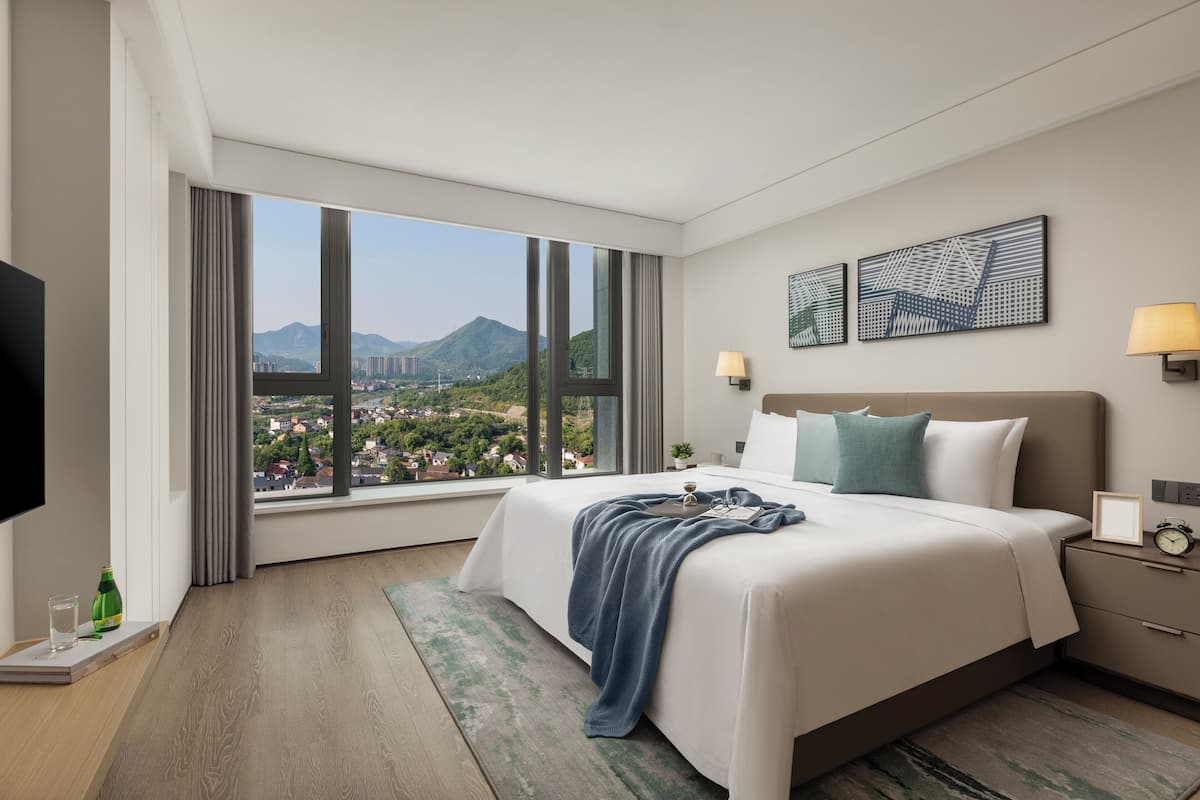 Ein Hotelzimmer mit großem Fenster und Blick auf die Stadt, einem Bett mit blauer Decke, einem Nachttisch mit Lampe und einem Flachbildfernseher.