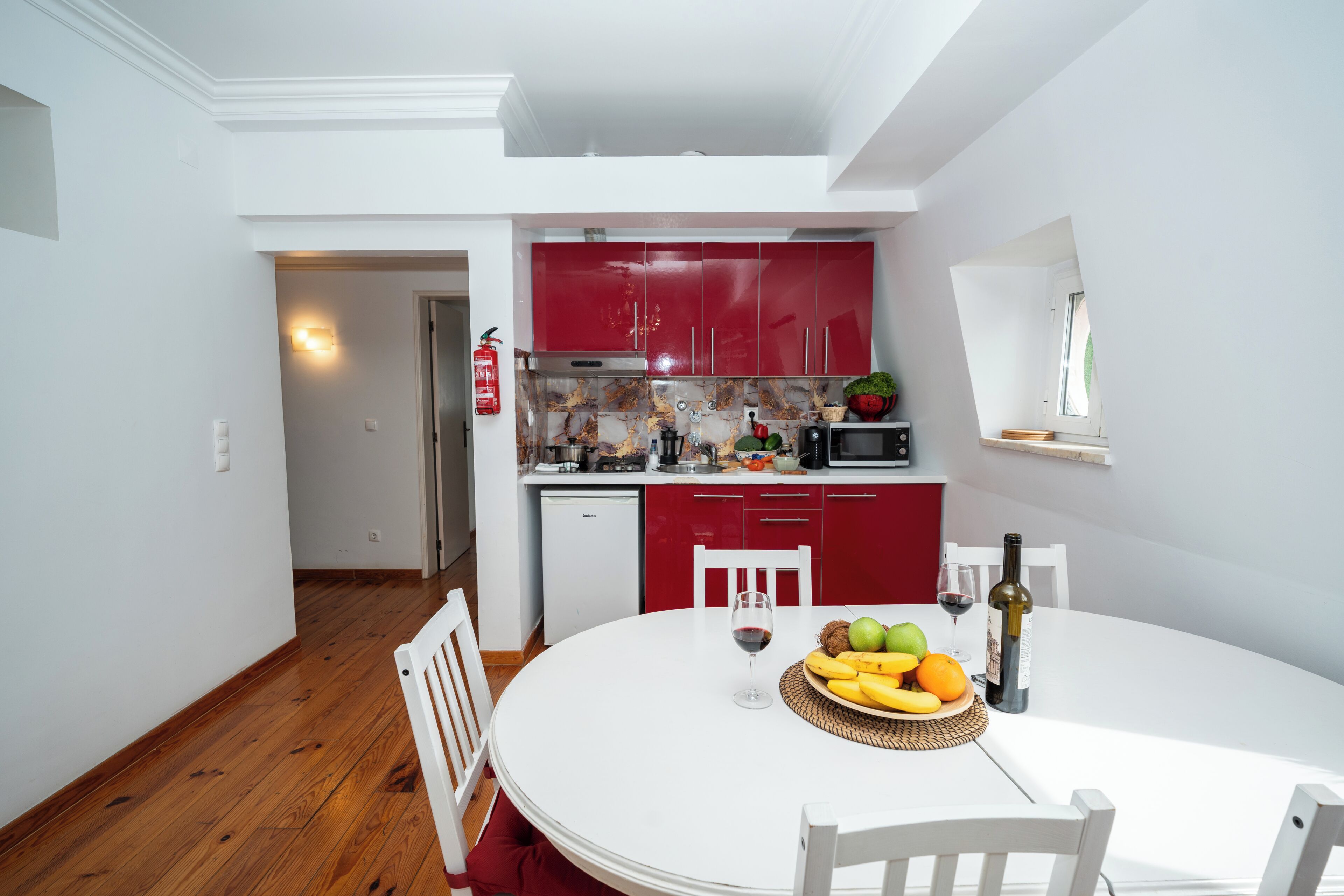 Una cocina moderna con armarios rojos, una mesa de comedor blanca con fruta y vino, y vistas al área de estar.